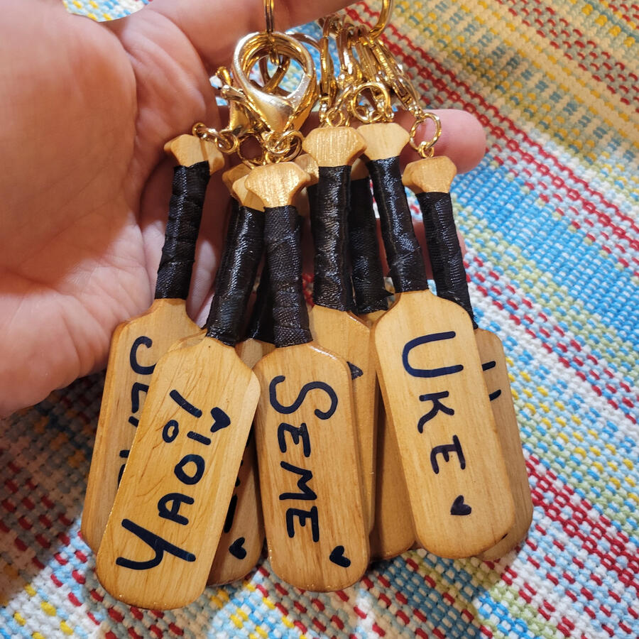 [V1] Wooden Yaoi Paddle Keychains; CNC cut pine with a dark pecan stain, black ribbon handle wraps, navy acrylic hand lettering, and rose gold hardware.